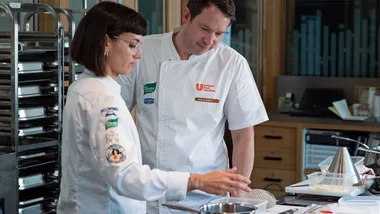 A male and female chef wearing their uniforms chatting in a kitchen